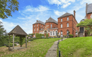 Property for sale by Auction A large, red-brick building with arched windows and a steep, gray-tiled roof sits against a bright blue sky. Manicured green lawns surround the structure, with a stone pathway leading up to it. Previously marketed by Palace Auctions, an open wooden gazebo stands in the foreground. Presented by Palace Auctions