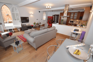 Property for sale by Auction A modern, open-concept living room and kitchen. The living area features two gray sofas, a TV mounted on the wall, and a red-themed painting. The kitchen has wooden cabinets, a central island, and stainless-steel appliances. A staircase leads to a bright doorway in this property previously marketed by Palace Auctions. Presented by Palace Auctions