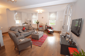 Property for sale by Auction A cozy living room with wooden flooring and white walls, featuring a gray sofa set with red and gray cushions, an ottoman, a red striped rug, a wall-mounted TV, and various decorative items including plants and framed photos. Two arched windows and a door provide natural light in this property previously marketed by Palace Auctions. Presented by Palace Auctions