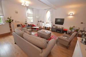 Property for sale by Auction A cozy living room with light wooden flooring features a gray sofa set facing a wall-mounted TV, a central ottoman, and a vibrant red rug. The space is illuminated by arched windows and decorated with plants and framed photos, creating a warm and inviting atmosphere in this property previously marketed by Palace Auctions. Presented by Palace Auctions