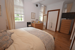 Property for sale by Auction A cozy bedroom, previously marketed by Palace Auctions, boasts a neatly made double bed with beige and blue bedding. The room features large windows with light curtains, a wooden dresser adorned with various items, a full-length mirror, a wardrobe, and a TV on a tall chest of drawers. Presented by Palace Auctions