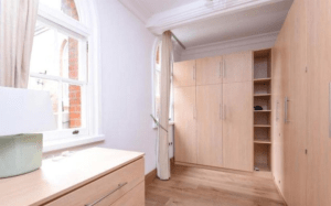 Property for sale by Auction A bright and tidy bedroom with light wood furniture, including a dresser on the left and built-in wardrobes on the right. Large arched windows with curtains let in ample natural light, illuminating the hardwood floor and white walls. This property was previously marketed by Palace Auctions. Presented by Palace Auctions