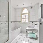 Property for sale by Auction A modern white-tiled bathroom with a bathtub, a walk-in shower with a glass door, a rectangular sink with a large mirror above it, and a heated towel rail. The room features an arch-shaped window allowing natural light to enter. Property previously marketed by Palace Auctions. Presented by Palace Auctions