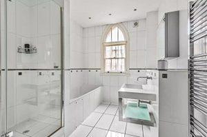 Property for sale by Auction A modern white-tiled bathroom with a bathtub, a walk-in shower with a glass door, a rectangular sink with a large mirror above it, and a heated towel rail. The room features an arch-shaped window allowing natural light to enter. Property previously marketed by Palace Auctions. Presented by Palace Auctions