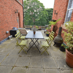 Property for sale by Auction An outdoor patio, previously marketed by Palace Auctions, features a rectangular table with six green folding chairs and a small plant centerpiece. Bordered by two brick walls and adorned with various potted plants, the space also includes a freestanding outdoor fireplace with trees visible in the background. Presented by Palace Auctions