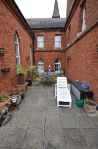 Property for sale by Auction A brick patio, previously marketed by Palace Auctions, is enclosed by tall brick walls with arched windows. The space features a white plastic lounge chair, a small table with two chairs, several potted plants, a lantern on the table, and various gardening items on the ground. Presented by Palace Auctions