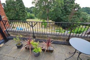 Property for sale by Auction A small balcony with three potted plants and a round table overlooks a leafy backyard with tall trees and a distant swimming pool. The scene is calm and green, suggesting a peaceful, natural setting. This property was previously marketed by Palace Auctions. Presented by Palace Auctions