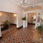 Property for sale by Auction A spacious, well-lit hallway with patterned tile flooring, white walls, and tall ceilings. A chandelier hangs from the ceiling, and there are plants in planters scattered throughout the space. Previously marketed by Palace Auctions, it features a large mirror and candle holder on a wooden table on the right. Presented by Palace Auctions