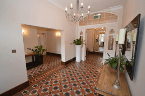 Property for sale by Auction A spacious, well-lit hallway with patterned tile flooring, white walls, and tall ceilings. A chandelier hangs from the ceiling, and there are plants in planters scattered throughout the space. Previously marketed by Palace Auctions, it features a large mirror and candle holder on a wooden table on the right. Presented by Palace Auctions