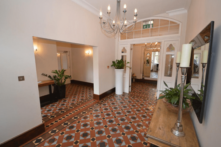 Property for sale by Auction A spacious, well-lit hallway with patterned tile flooring, white walls, and tall ceilings. A chandelier hangs from the ceiling, and there are plants in planters scattered throughout the space. Previously marketed by Palace Auctions, it features a large mirror and candle holder on a wooden table on the right. Presented by Palace Auctions