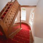 Property for sale by Auction A wooden staircase with intricate detailing on the bannister and red carpet with a patterned design ascends upwards. Three arched windows with stained glass are visible on the landing, allowing light to filter through. The property, previously marketed by Palace Auctions, features walls painted white. Presented by Palace Auctions
