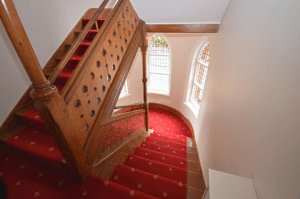 Property for sale by Auction A wooden staircase with intricate detailing on the bannister and red carpet with a patterned design ascends upwards. Three arched windows with stained glass are visible on the landing, allowing light to filter through. The property, previously marketed by Palace Auctions, features walls painted white. Presented by Palace Auctions