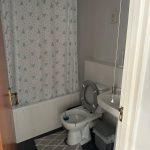 Property for sale by Auction A bathroom in Staines-upon-Thames with white walls and a dark floor is shown. It features a bathtub with a patterned shower curtain, a toilet with the lid up, a sink with a glass on the edge, and a small waste bin. Two matching rugs are placed on the floor of this cozy part of 13 Maynard Court. Presented by Palace Auctions