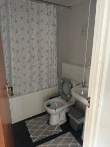 Property for sale by Auction A bathroom in Staines-upon-Thames with white walls and a dark floor is shown. It features a bathtub with a patterned shower curtain, a toilet with the lid up, a sink with a glass on the edge, and a small waste bin. Two matching rugs are placed on the floor of this cozy part of 13 Maynard Court. Presented by Palace Auctions