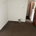 Property for sale by Auction A small, empty room in Staines-upon-Thames with white walls and a brown carpet. There is a grey lamp on the floor near the wall on the right side. An open door reveals a hallway with framed pictures on the walls and a partially visible radiator. A round white ceiling light is centered above, part of 13 Maynard Court, TW18 4QD. Presented by Palace Auctions