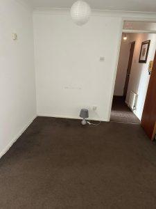 Property for sale by Auction A small, empty room in Staines-upon-Thames with white walls and a brown carpet. There is a grey lamp on the floor near the wall on the right side. An open door reveals a hallway with framed pictures on the walls and a partially visible radiator. A round white ceiling light is centered above, part of 13 Maynard Court, TW18 4QD. Presented by Palace Auctions