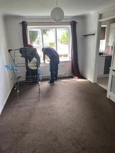 Property for sale by Auction A person stands by a window in a mostly empty room at part of 13 Maynard Court, Rosefield Road, TW18 4QD, adjusting or cleaning the window. A laundry drying rack with clothes is on the left side near the window. Dark curtains frame the window, and a kitchen area is visible to the right. Presented by Palace Auctions