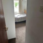 Property for sale by Auction View of a small bedroom through a slightly open door, part of 13 Maynard Court. The room has a single bed with a light gray blanket, and there is a window with light curtains allowing natural light to enter. The floor is carpeted, and a light switch is visible on the right side of the door frame. Presented by Palace Auctions