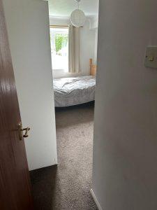 Property for sale by Auction View of a small bedroom through a slightly open door, part of 13 Maynard Court. The room has a single bed with a light gray blanket, and there is a window with light curtains allowing natural light to enter. The floor is carpeted, and a light switch is visible on the right side of the door frame. Presented by Palace Auctions