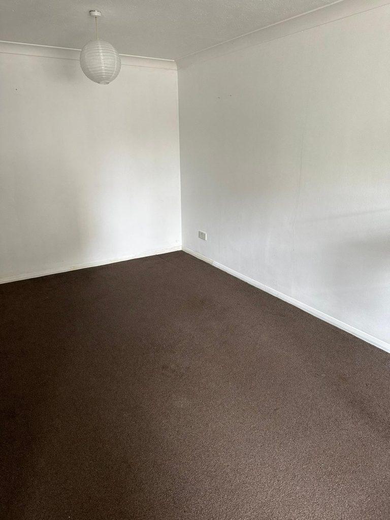 Property for sale by Auction An empty, unadorned room with plain white walls, a white ceiling, and a brown carpet. A single, round white light fixture hangs from the ceiling. The room has no furniture or decorations and is part of 13 Maynard Court on Rosefield Road in Staines-upon-Thames. Presented by Palace Auctions