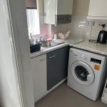 Property for sale by Auction A cozy kitchen corner on Rosefield Road, TW18 4QD, Staines-upon-Thames features a sink and drying rack on the left, a countertop with a paper towel holder and an electric kettle, and a washing machine below. The cabinets above and below are a mix of white and gray, with a partially visible window. Presented by Palace Auctions