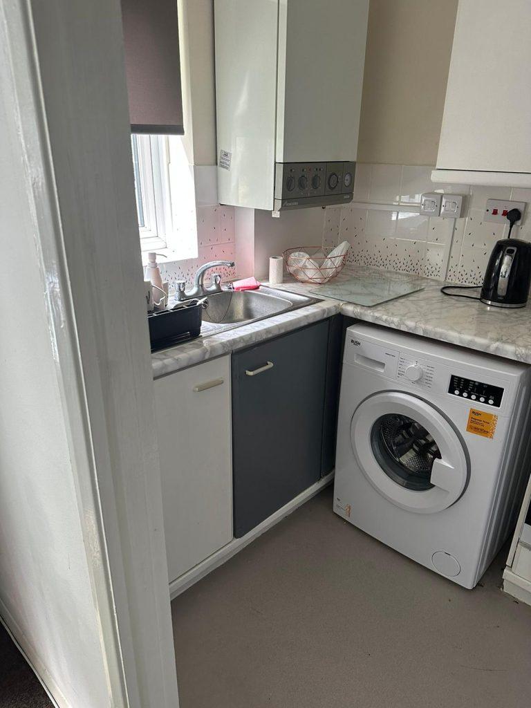 Property for sale by Auction A cozy kitchen corner on Rosefield Road, TW18 4QD, Staines-upon-Thames features a sink and drying rack on the left, a countertop with a paper towel holder and an electric kettle, and a washing machine below. The cabinets above and below are a mix of white and gray, with a partially visible window. Presented by Palace Auctions
