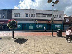 Property for sale by Auction A commercial building in Crewe, Cheshire, with turquoise boards stating "SORRY! WE HAVE CLOSED," spans the width of the former Poundland sold by Palace Auctions. The surrounding area features a hanging flower basket, lamp posts, and a few pedestrians walking nearby. The sky above is partly cloudy. Presented by Palace Auctions