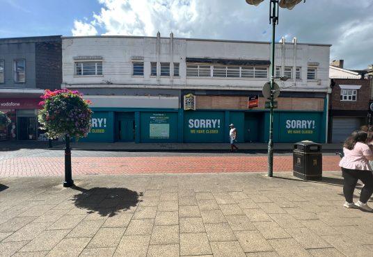 Property for sale by Auction A commercial building in Crewe, Cheshire, with turquoise boards stating "SORRY! WE HAVE CLOSED," spans the width of the former Poundland sold by Palace Auctions. The surrounding area features a hanging flower basket, lamp posts, and a few pedestrians walking nearby. The sky above is partly cloudy. Presented by Palace Auctions