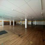 Property for sale by Auction A large, empty indoor space in a former Poundland in Crewe, Cheshire, with a wooden floor and white ceiling panels supported by numerous pillars. The walls feature sections of white, red, and yellow paint. Scattered dark patches mark the floor, and a few overhead fluorescent lights illuminate the area. Presented by Palace Auctions