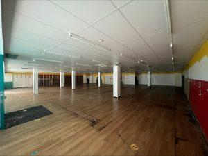 Property for sale by Auction A large, empty indoor space in a former Poundland in Crewe, Cheshire, with a wooden floor and white ceiling panels supported by numerous pillars. The walls feature sections of white, red, and yellow paint. Scattered dark patches mark the floor, and a few overhead fluorescent lights illuminate the area. Presented by Palace Auctions