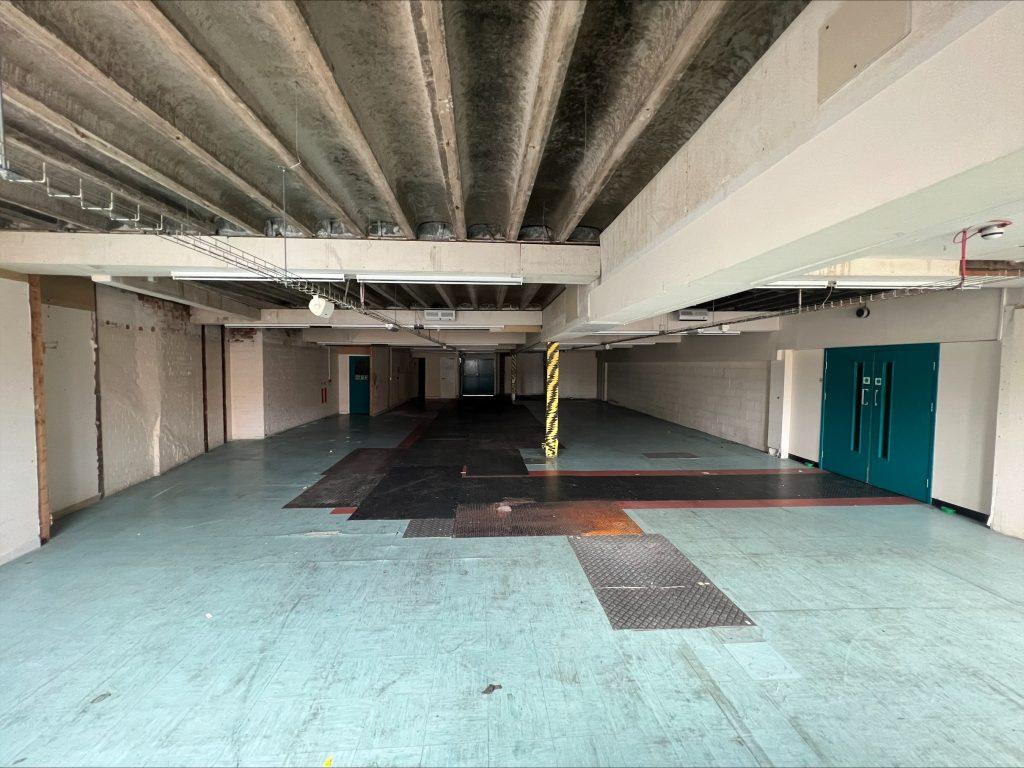 Property for sale by Auction A large, dimly lit, empty industrial space with a high, unfinished ceiling in a commercial building in Crewe Cheshire. The floor has a mix of blue and black tiled sections, with some areas appearing worn out. Walls are plain and partially covered in patches of exposed brick. Two large blue doors are visible on the right. Presented by Palace Auctions