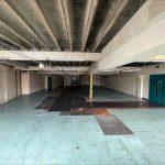 Property for sale by Auction A large, dimly lit, empty industrial space with a high, unfinished ceiling in a commercial building in Crewe Cheshire. The floor has a mix of blue and black tiled sections, with some areas appearing worn out. Walls are plain and partially covered in patches of exposed brick. Two large blue doors are visible on the right. Presented by Palace Auctions