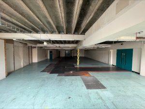 Property for sale by Auction A large, dimly lit, empty industrial space with a high, unfinished ceiling in a commercial building in Crewe Cheshire. The floor has a mix of blue and black tiled sections, with some areas appearing worn out. Walls are plain and partially covered in patches of exposed brick. Two large blue doors are visible on the right. Presented by Palace Auctions