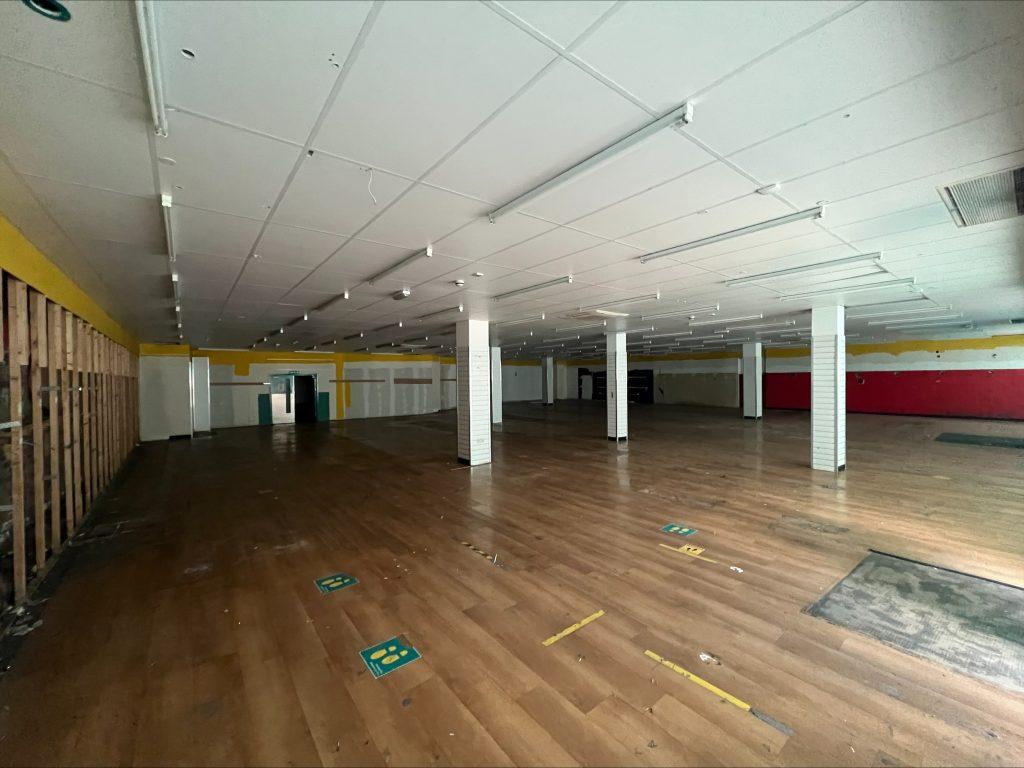 Property for sale by Auction A large, empty room with a wooden floor and white ceiling tiles sits within a former Poundland in Crewe, Cheshire. The space is illuminated by natural light from hidden windows, featuring several white columns and noticeable green markings on the floor. The walls are vibrantly yellow and red. Presented by Palace Auctions