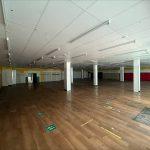 Property for sale by Auction A large, empty room with a wooden floor and white ceiling tiles sits within a former Poundland in Crewe, Cheshire. The space is illuminated by natural light from hidden windows, featuring several white columns and noticeable green markings on the floor. The walls are vibrantly yellow and red. Presented by Palace Auctions