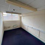 Property for sale by Auction A small, empty room with white brick walls, a single window letting in natural light, a navy blue carpet, and a ceiling with fluorescent lighting. Located in a commercial building in Crewe Cheshire, this former Poundland sold by Palace Auctions features visible electrical conduits and outlets needing maintenance. Presented by Palace Auctions