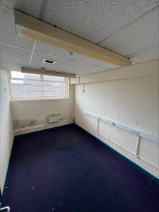 Property for sale by Auction A small, empty room with white brick walls, a single window letting in natural light, a navy blue carpet, and a ceiling with fluorescent lighting. Located in a commercial building in Crewe Cheshire, this former Poundland sold by Palace Auctions features visible electrical conduits and outlets needing maintenance. Presented by Palace Auctions