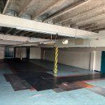 Property for sale by Auction A large, empty indoor space with concrete ceilings and partially tiled flooring. Blue double doors are visible on the right side, while the walls are mostly white with some tiled sections. Metal support beams and pipes are exposed, and the room is well-lit from above—a former Poundland in a commercial building in Crewe Cheshire sold by Palace Auctions. Presented by Palace Auctions