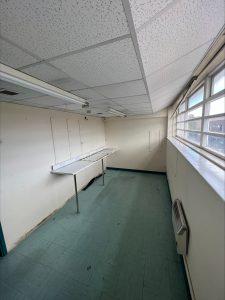 Property for sale by Auction A small, empty room in a commercial building in Crewe Cheshire with a tiled ceiling and green linoleum floor. The room features a long, narrow table attached to the left wall, several wall-mounted electrical outlets, a large window on the right side, and a wall-mounted heater below the window. Presented by Palace Auctions