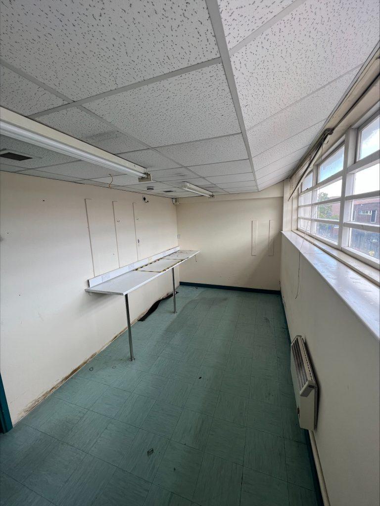 Property for sale by Auction A small, empty room in a commercial building in Crewe Cheshire with a tiled ceiling and green linoleum floor. The room features a long, narrow table attached to the left wall, several wall-mounted electrical outlets, a large window on the right side, and a wall-mounted heater below the window. Presented by Palace Auctions