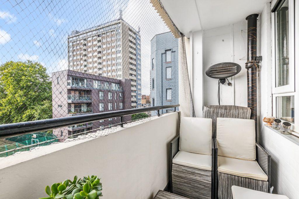 A small balcony at Flat 3, 1 Macclesfield House, Central Street, LONDON EC1V 8BA features a wicker chair with beige cushions and is surrounded by a safety net. Potted plants grace the table in the foreground as sunlight filters in, illuminating nearby buildings, trees, and a blue sky. Ideal investment for sale by auction. Presented by Palace Auctions