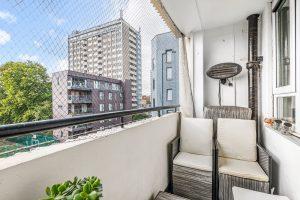 A small balcony at Flat 3, 1 Macclesfield House, Central Street, LONDON EC1V 8BA features a wicker chair with beige cushions and is surrounded by a safety net. Potted plants grace the table in the foreground as sunlight filters in, illuminating nearby buildings, trees, and a blue sky. Ideal investment for sale by auction. Presented by Palace Auctions