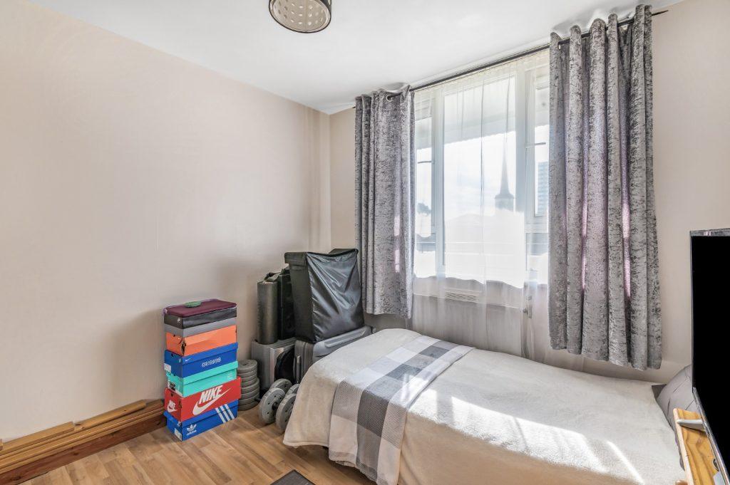 A small, tidy bedroom at Flat 3,1 Macclesfield House Central Street LONDON EC1V 8BA features light brown wood flooring and a single bed covered with a white and gray blanket. The window has sheer curtains and darker drapes. Stacks of colorful shoe boxes and a suitcase are placed beside the bed as sunlight filters through. This investment is for sale by auction. Presented by Palace Auctions