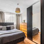 Modern bedroom featuring a neatly made bed with grey bedding, framed by a tufted headboard. A wooden dresser with a potted plant atop it stands against the wall, opposite a large mirrored wardrobe. Curtains cover the window, and a ceiling light hangs above. Flat 3,1 Macclesfield House Central Street LONDON EC1V 8BA investment for sale by auction. Presented by Palace Auctions