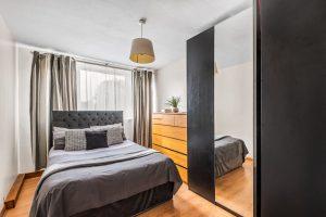 Modern bedroom featuring a neatly made bed with grey bedding, framed by a tufted headboard. A wooden dresser with a potted plant atop it stands against the wall, opposite a large mirrored wardrobe. Curtains cover the window, and a ceiling light hangs above. Flat 3,1 Macclesfield House Central Street LONDON EC1V 8BA investment for sale by auction. Presented by Palace Auctions