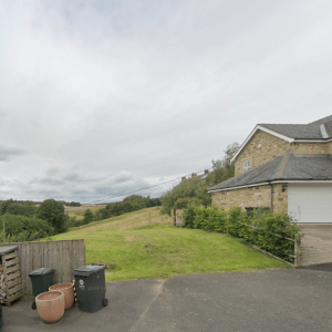 Property for sale by Auction A countryside scene showing part of a residential house with a stone exterior and a garage. The house is surrounded by greenery and a spacious lawn that extends to open fields. In the foreground are a wooden fence, two plant pots, and a couple of trash bins. Presented by Palace Auctions