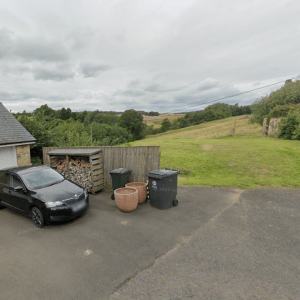 Property for sale by Auction A black car is parked on a driveway next to a building with a wooden log storage area and three large pots nearby. Two trash bins are also present. The background showcases a grassy yard, rolling hills, and a cloudy sky. Presented by Palace Auctions