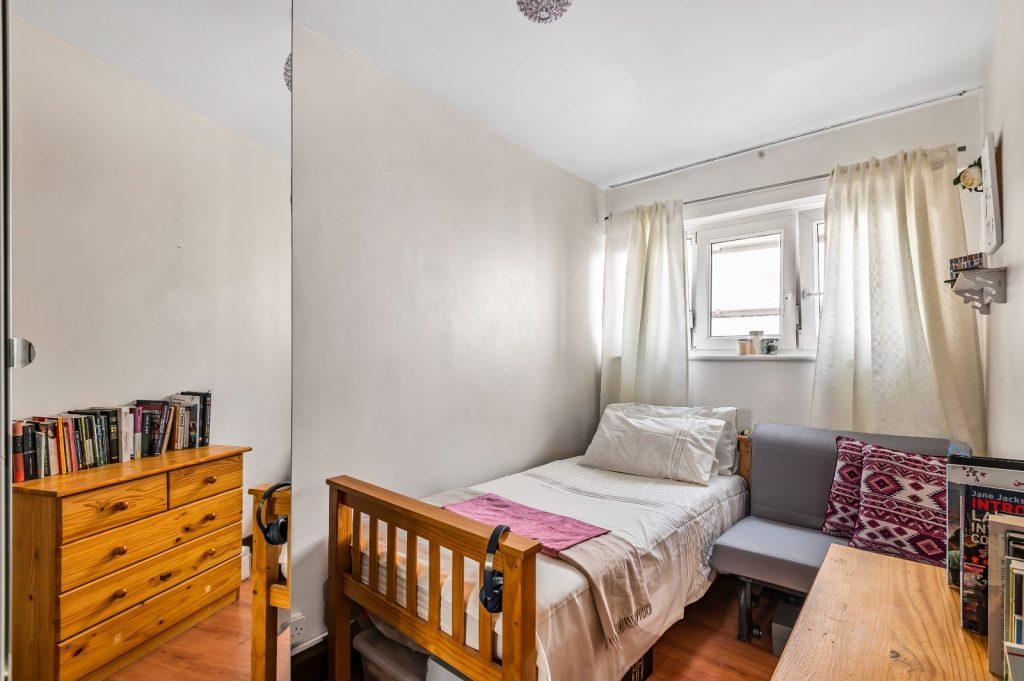 Flat 3 at Macclesfield House, Central Street, LONDON EC1V 8BA features a small, cozy bedroom with a single bed adorned with a white and pink blanket. Beside the bed sits a grey chair and a light wooden chest of drawers topped with books. Natural light filters through the window with white curtains, highlighting the room's light-colored walls and hardwood flooring. This investment Presented by Palace Auctions