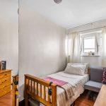 Flat 3 at Macclesfield House, Central Street, LONDON EC1V 8BA features a small, cozy bedroom with a single bed adorned with a white and pink blanket. Beside the bed sits a grey chair and a light wooden chest of drawers topped with books. Natural light filters through the window with white curtains, highlighting the room's light-colored walls and hardwood flooring. This investment Presented by Palace Auctions