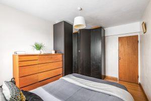 A cozy bedroom in Flat 3, 1 Macclesfield House, Central Street LONDON EC1V 8BA features a large wooden dresser with a potted plant and books on top. The bed is neatly made with grey and white bedding. Black wardrobes occupy a corner, and a wooden door leads out of the room. A white ceiling light fixture hangs above. Presented by Palace Auctions
